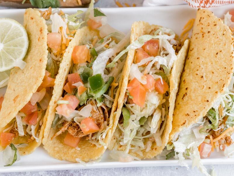 Four soft tacos on a plate topped with shredded chicken, salsa and shredded lettuce.
