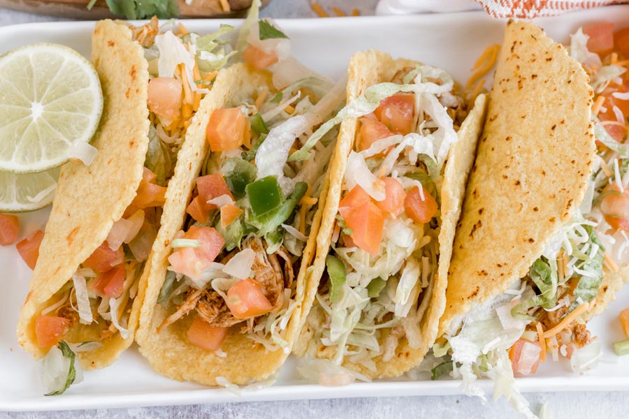 Four soft tacos on a plate topped with shredded chicken, salsa and shredded lettuce.