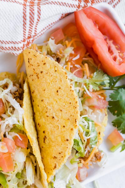 Two tacos filled with pico de gallo, cheese and shredded lettuce with a big tomato at the top of the plate.