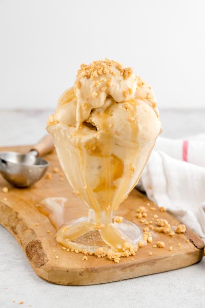Tall serving of creamy keto Twix ice cream with caramel sauce and cookie crumbles overflowing from a glass sundae dish.