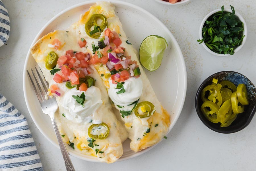 Two enchiladas on a plate topped with cheese, pico de gallo and a white sauce. To the plate is a bowl of jalapenos and cilantro.