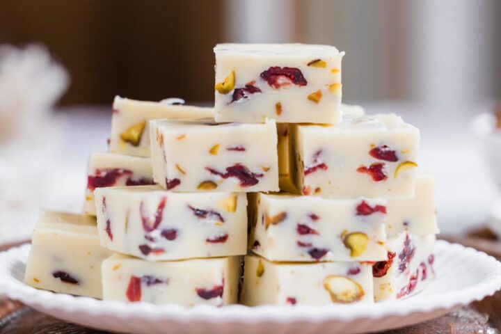 A stack of creamy white chocolate fudge squares studded with cranberries and pistachios sits on a white plate.