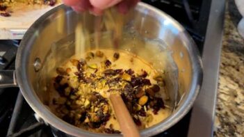 Chopped pistachios and dried cranberries are added into melted white chocolate in a saucepan.