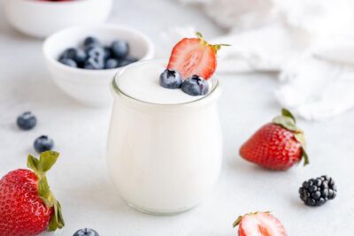 A plain yogurt in a jar with two blueberries and a sliced strawberry inside. Berries around surrounding the jar.