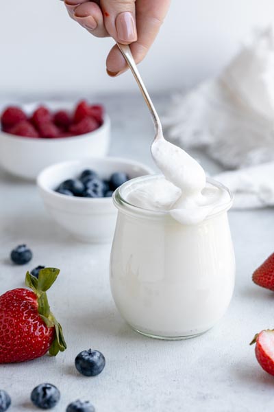 A hand holding a spoonful of white yogurt in front of the jar of yogurt. Some of the yogurt is spilling down the edge of the glass.