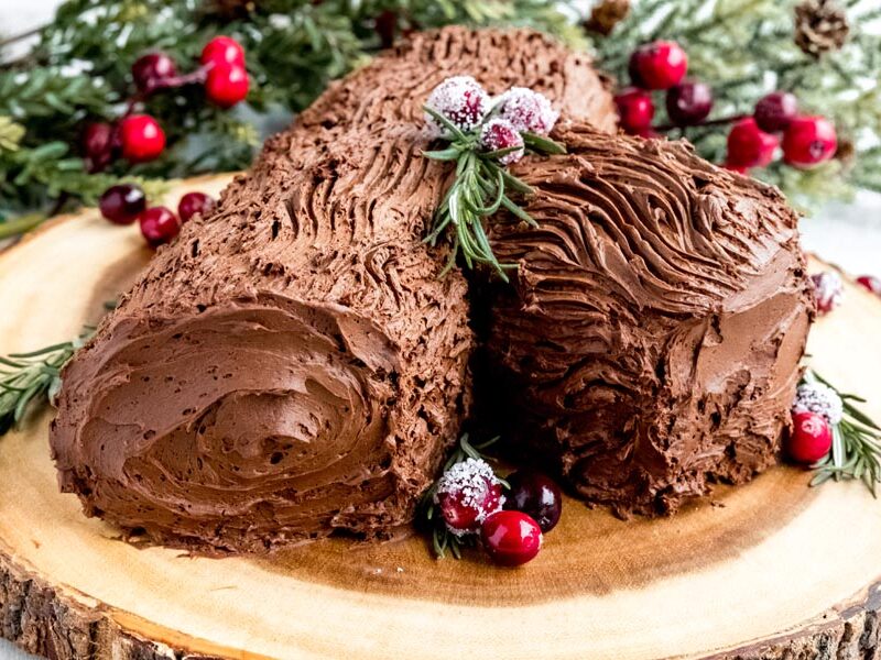 A chocolate yule log cake decorated with cranberries and rosemary on a wooden board.