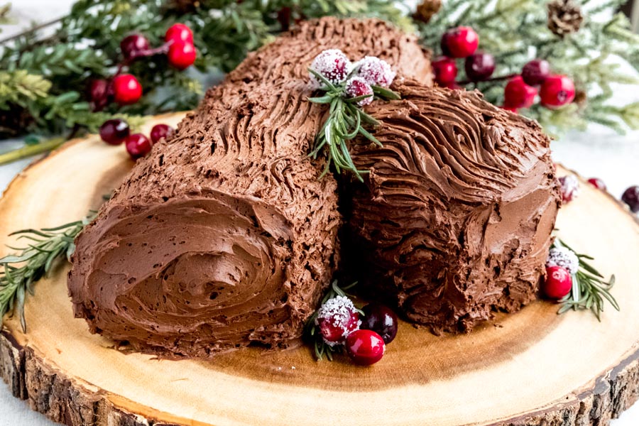 A chocolate yule log cake decorated with cranberries and rosemary on a wooden board.