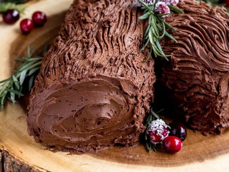 Dark chocolate ganache frosting decorated to look like bark and a log next to rosemary sprigs and cranberries.