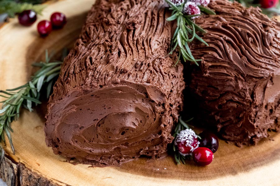 Dark chocolate ganache frosting decorated to look like bark and a log next to rosemary sprigs and cranberries.