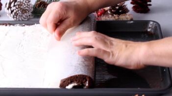 Hands rolling the thin cake using parchment paper to help roll tightly.