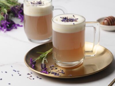 Two mugs of warm lavender milk tea topped with creamy foam and dried culinary lavender, styled with fresh lavender and honey.