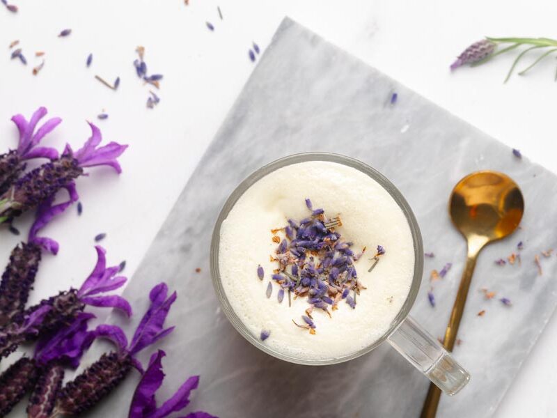 Overhead view of frothy lavender milk tea garnished with dried lavender buds beside fresh purple lavender flowers.