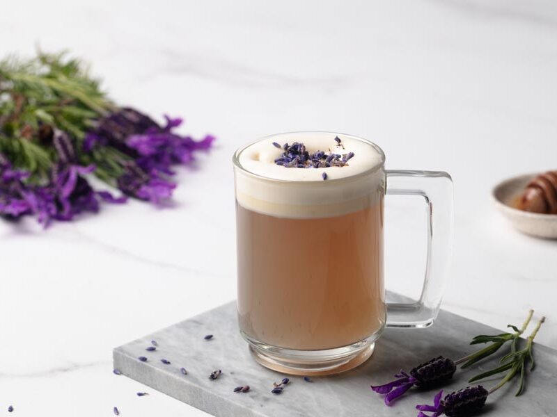 A warm mug of lavender milk tea topped with milk foam and dried lavender buds on a marble serving board.