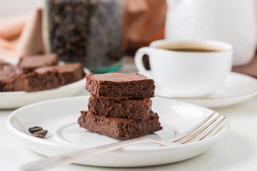 A stack of three rich chocolate brownies sits on a white plate next to a coffee cup and forks.