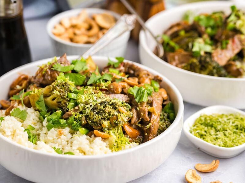 Two bowls of broccoli and cashew beef over cauliflower rice with lime zest nearby.
