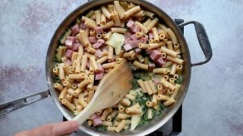Cooked pasta and diced ham being mixed into the creamy spinach sauce.