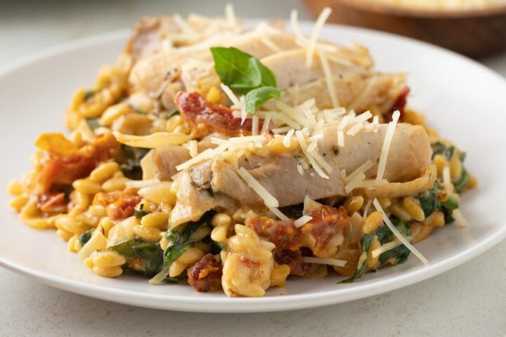 A plate with creamy orzo pasta with spinach and sun dried tomatoes topped with sliced chicken and shredded parmesan cheese.