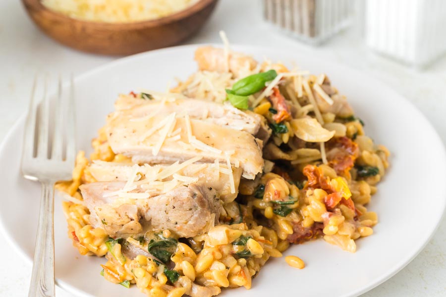 A plate of cooked chicken over a bed of creamy pasta filled with tomato and spinach.