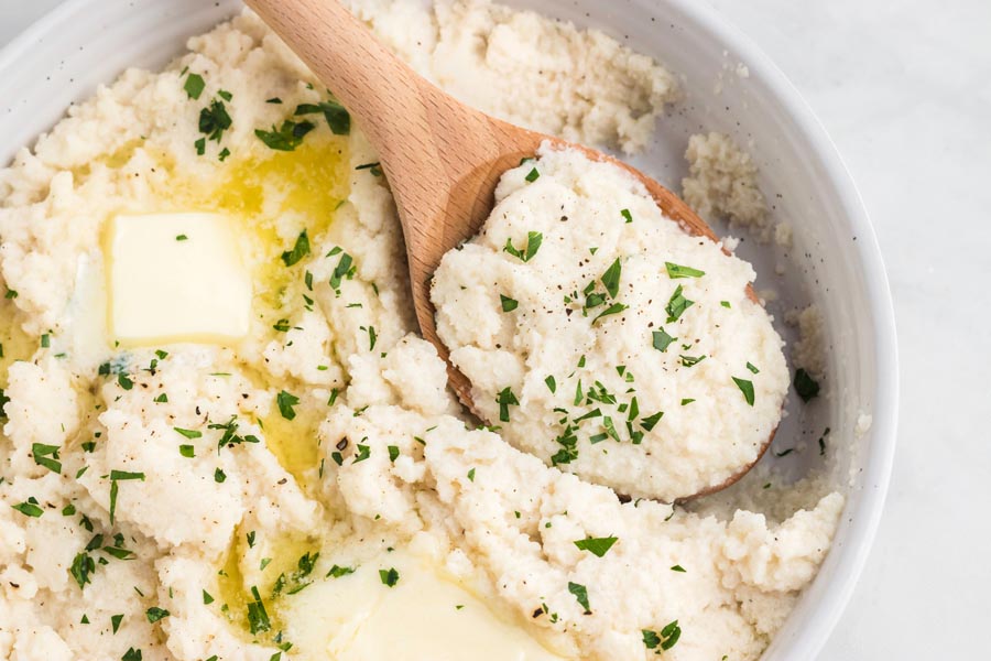 A wooden spoon digging into a bowl of mashed turnips topped with a cube of melting butter and parsley.