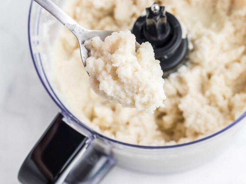 A spoon filled with mashed turnips over a food processor.