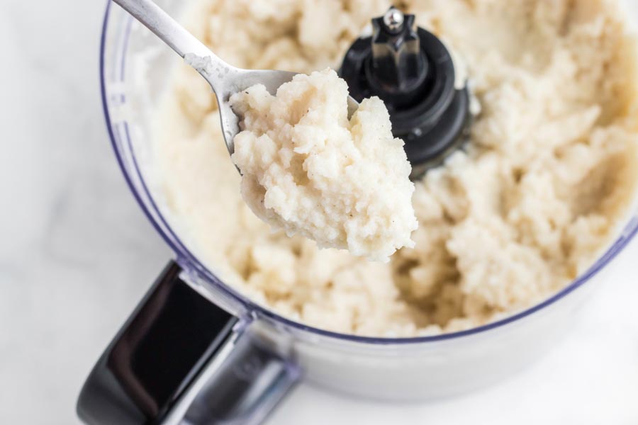 A spoon filled with mashed turnips over a food processor.