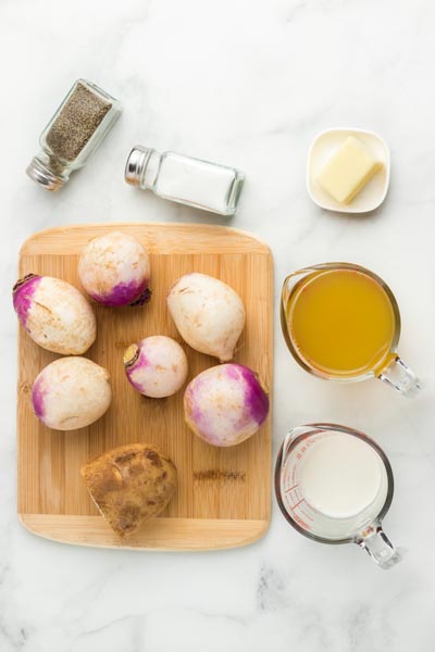 Whole turnips on a cutting board with a half a potato, butter, salt and pepper, cream and chicken broth.