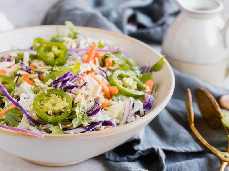 A bowl with coleslaw, jalapeno slices, carrots and red onion with dressing nearby.