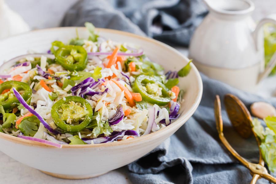 A bowl with coleslaw, jalapeno slices, carrots and red onion with dressing nearby.