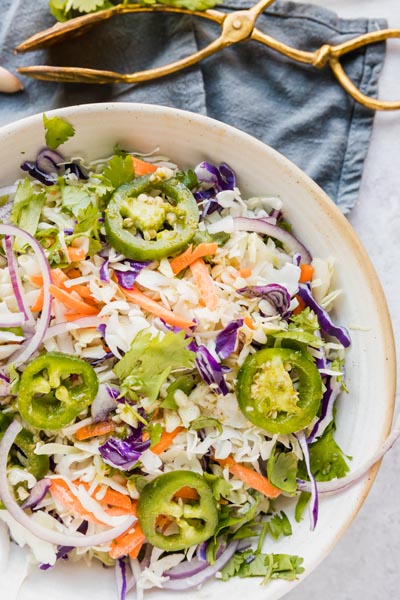 A large bowl of coleslaw topped with jalapeno slices near some gold tongs.