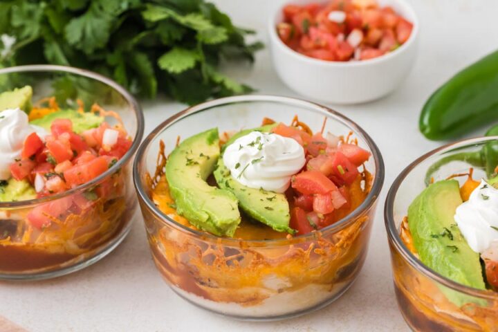Individual Mexican lasagna bowls topped with avocado, pico de gallo, and sour cream in glass containers.