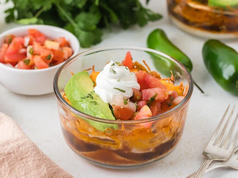 A close-up of a single Mexican lasagna bowl surrounded by fresh jalapeños, pico de gallo, and herbs.