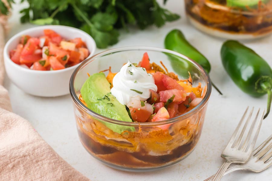 A close-up of a single Mexican lasagna bowl surrounded by fresh jalapeños, pico de gallo, and herbs.