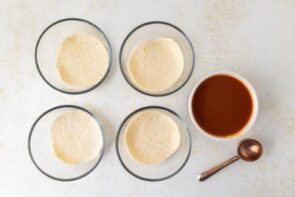 Step showing tortillas placed in the bottom of each bowl next to a dish of enchilada sauce.