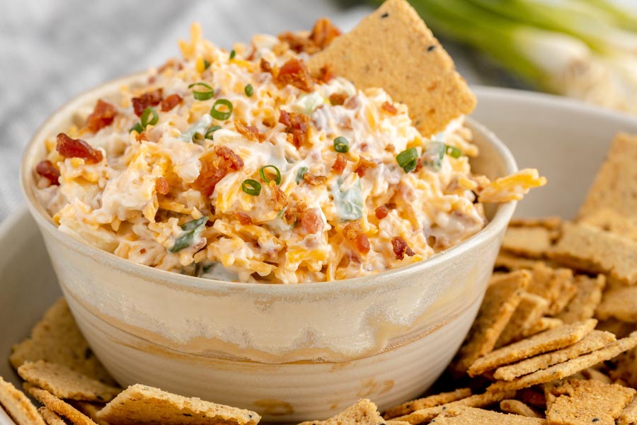 Creamy dip with bacon, green onion and cheese in a bowl next to a pile of crackers