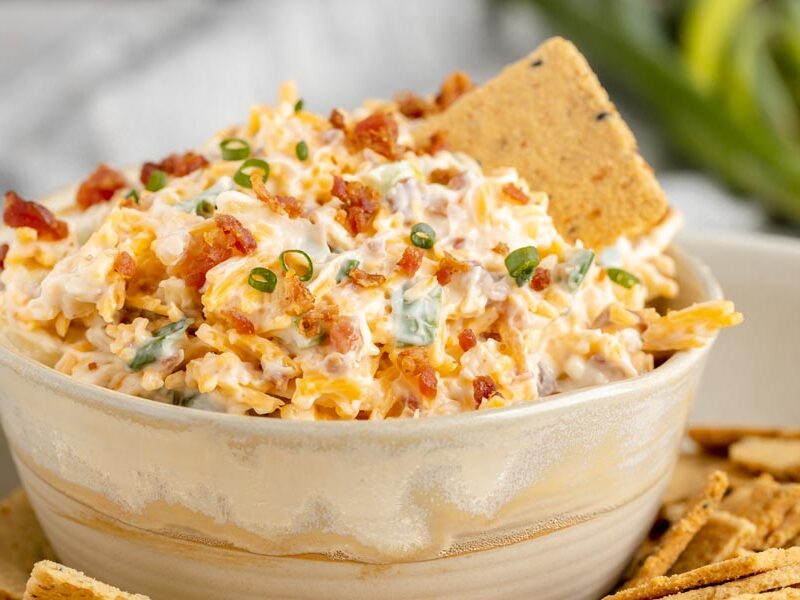 a creamy dip filled with bacon bits, cheddar cheese and green onion with a cracker stuck in the dip