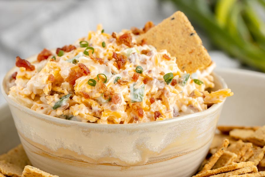 a creamy dip filled with bacon bits, cheddar cheese and green onion with a cracker stuck in the dip