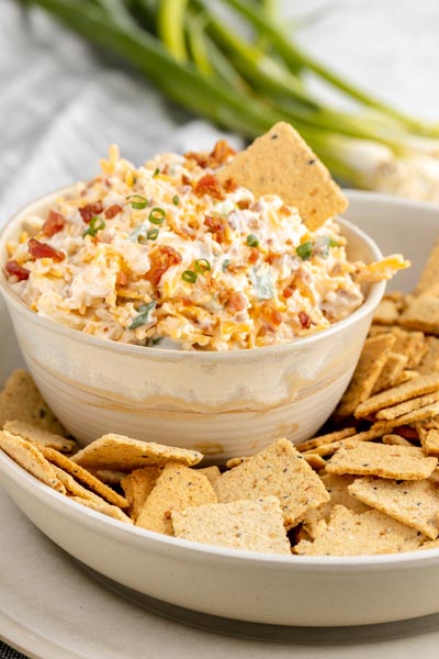 a plate of crackers next to bowl of dip topped with bacon and green onions in the background