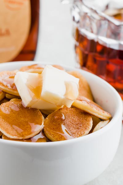 mini pancakes in a bowl topped with two cubes of butter and a drizzle of syrup