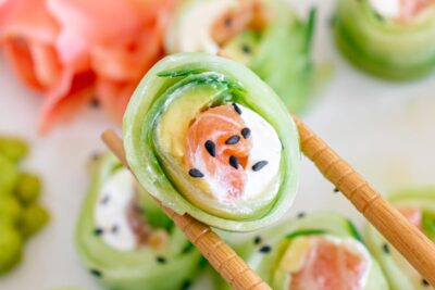 Chopsticks holding a cucumber wrapped sushi roll filled with salmon, cream cheese and avocado and topped with black sesame seeds.