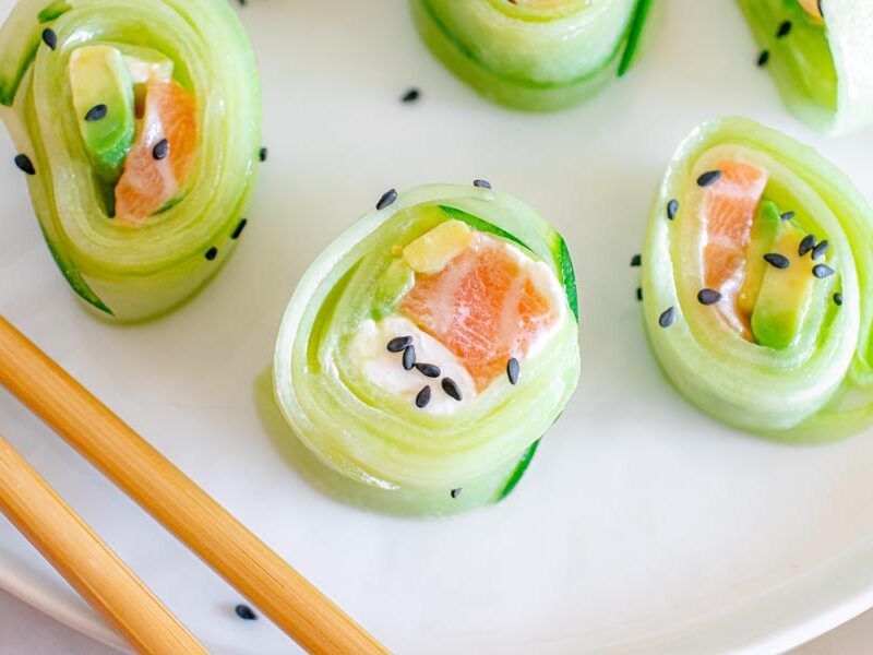 Cucumber wrapped sushi rolls on a plate next to chopsticks and topped with sesame seeds.