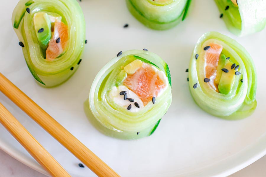 Cucumber wrapped sushi rolls on a plate next to chopsticks and topped with sesame seeds.