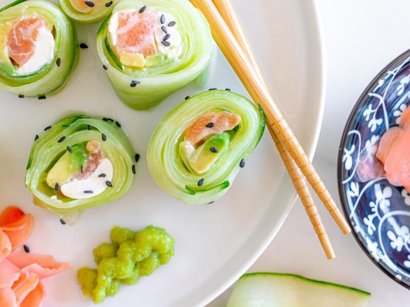 Cucumber rolls on a white plate next to wasabi and pickled ginger. Sushi is filled with salmon, cream cheese and avocado.