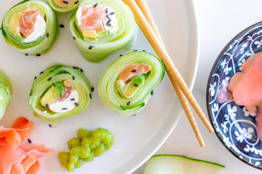 Cucumber rolls on a white plate next to wasabi and pickled ginger. Sushi is filled with salmon, cream cheese and avocado.
