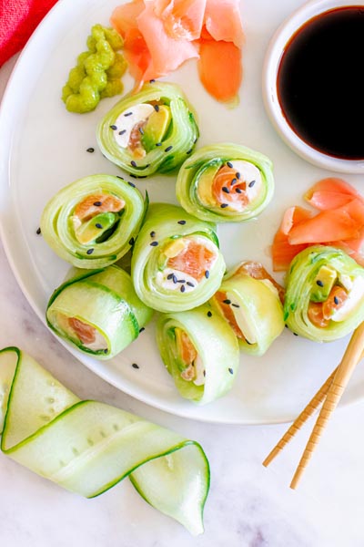 A plate of naruto rolls sushi wrapped in cucumber and filled with salmon and cream cheese. On the plate there is also a bowl of soy sauce, pickled ginger and green wasabi paste.