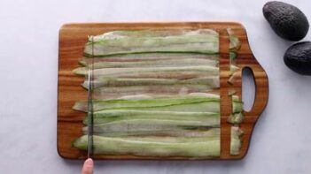 Long strips of cucumber on a wooden cutting board. A knife cutting the ends off the cucumbers to make them even.