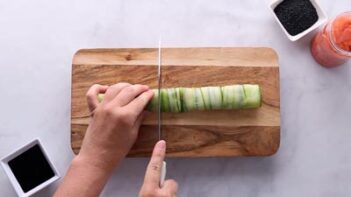 Cutting a cucumber wrapped sushi roll with a knife on a wooden cutting board.