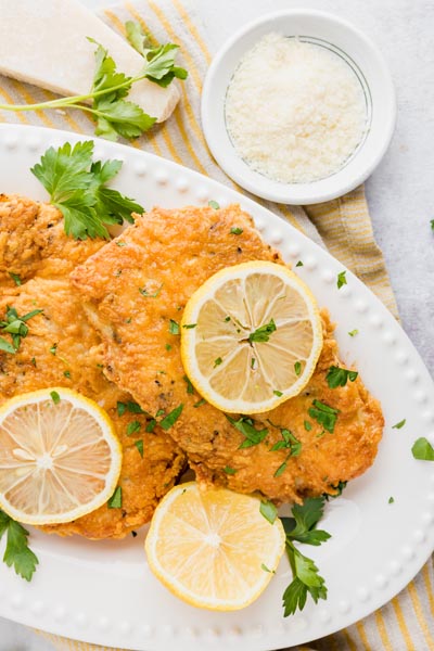 a plate of parmesan crusted chicken covered in lemon wedges and chopped parsley