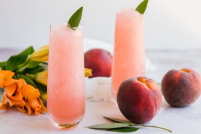 two blended peach cocktails with fresh peaches near by and orange flowers
