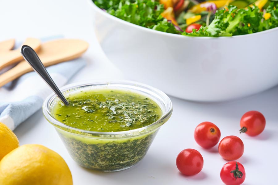 A small bowl of pesto dressing containing basil with a spoon inside next to grape tomatoes and a bowl of salad.