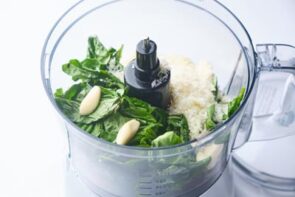 A food processor with fresh basil, garlic and grated parmesan cheese inside.